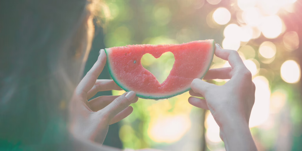 Woman holding a watermelon slice with a heart-shaped cutout in natural sunlight.